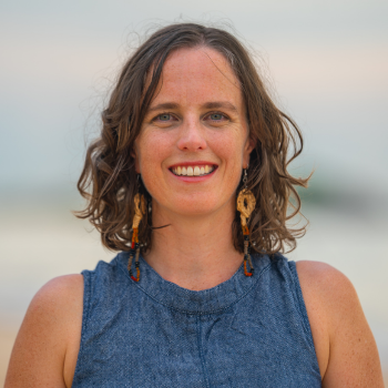 Outdoor headshot of a smiling woman with short curly hair, wearing a sleeveless blue top and long beaded earrings, with a soft blurred background.